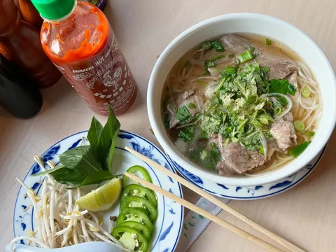 Bowl of beef phở with rice noodles, sliced beef, cilantro and scallions in a blue-and-white bowl at Pho Lang Thang, with a side plate of bean sprouts, Thai basil, lime and sliced jalapeños