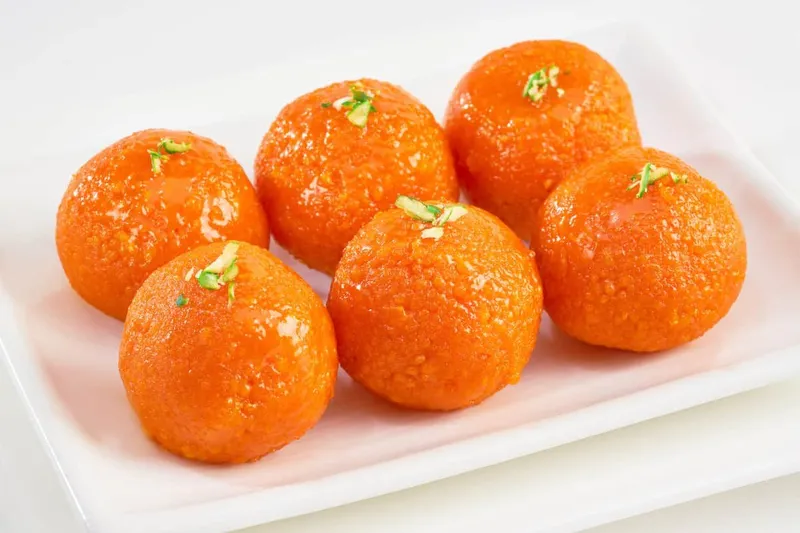 Six bright orange motichoor laddoo, ghee-fried gram-flour sweet balls topped with slivered pistachio, on a white plate, from Mithaas in Edison, New Jersey
