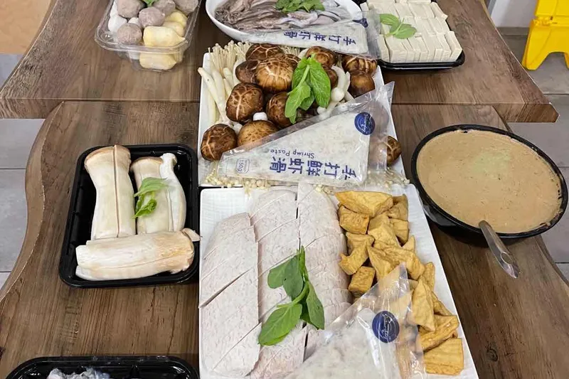 Overhead spread of fresh hot pot ingredients — mushrooms, tofu, sliced meats, prawns, scallops and shrimp paste — on a wood table at Xi'an Noodles