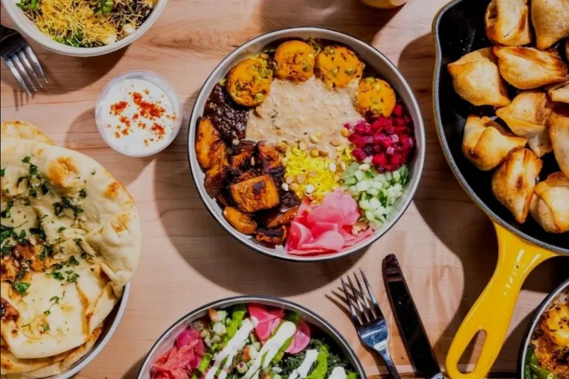 Overhead spread of RASA bowls, naan, samosas, raita, and colorful drinks on a wood table