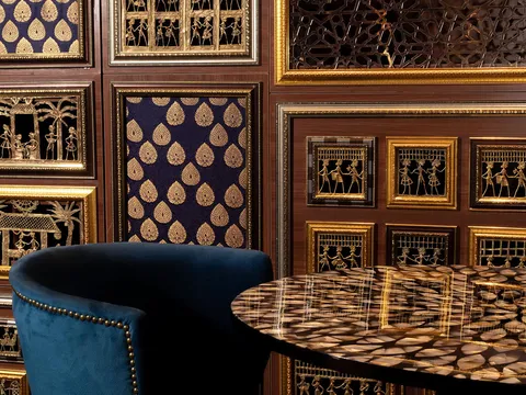Ornately paneled dining room at Musaafer in Houston, with a blue velvet chair and an inlaid stone table against walls of framed gold metalwork and brocade