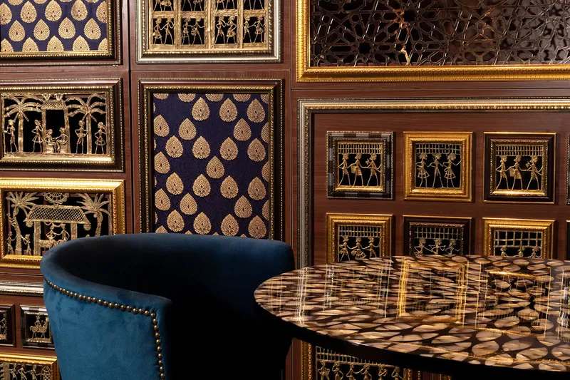Ornately paneled dining room at Musaafer in Houston, with a blue velvet chair and an inlaid stone table against walls of framed gold metalwork and brocade