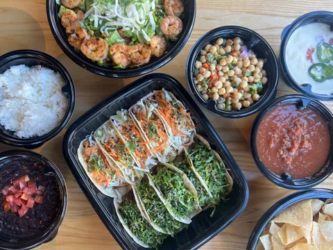 Overhead spread of Taqueria Tsunami fusion tacos, a grilled shrimp bowl, rice, black beans, chickpea salad, queso, salsa and tortilla chips in black takeout containers