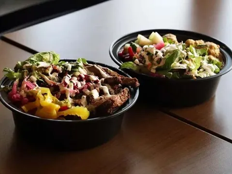 Two Indian rice bowls with grilled meat, greens, pickled onion, and drizzled sauces on a wooden table at The Indian Belly in Olathe