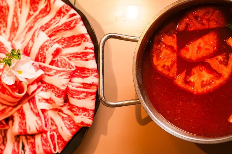 Top-down view of a marbled beef platter beside a tomato-broth hot pot at Little Sheep Hot Pot & Bar
