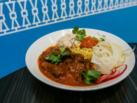A Bhuna curry bowl — braised meat curry over turmeric basmati rice with raita, mango pickle, shaved fennel and radish — on the restaurant's signature blue backdrop