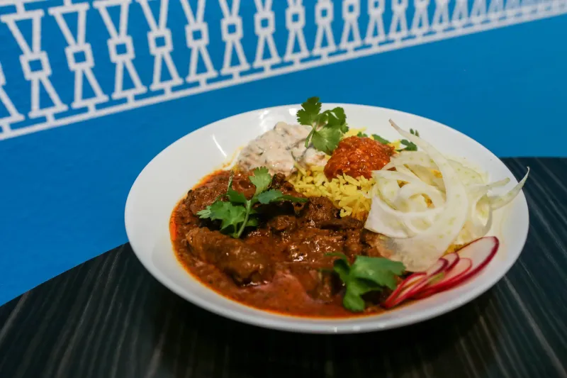 A Bhuna curry bowl — braised meat curry over turmeric basmati rice with raita, mango pickle, shaved fennel and radish — on the restaurant's signature blue backdrop