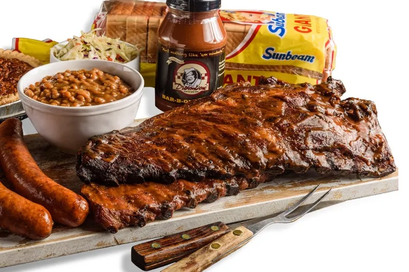 Dreamland Bar-B-Que full slab of ribs on a cutting board with two smoked sausage links, a bowl of baked beans, coleslaw, a pecan pie, a bottle of Dreamland BBQ sauce and Sunbeam bread