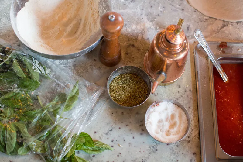 Il Lazzarone pizza-making ingredients on a marble counter — flour, oregano, sea salt, San Marzano tomato sauce, fresh basil and a copper olive-oil pourer