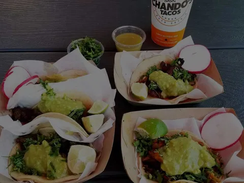 Overhead view of Chando's Tacos street tacos in paper boats with lime, radish, and salsa, alongside a branded Chando's Tacos cup