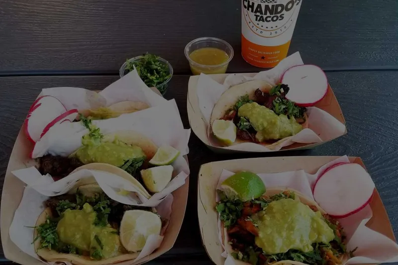 Overhead view of Chando's Tacos street tacos in paper boats with lime, radish, and salsa, alongside a branded Chando's Tacos cup