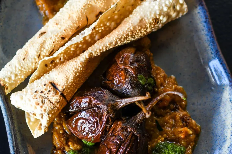 Baby Brinjals — roasted stuffed baby eggplants over spiced masala with green chutney and rolled papadum on a blue ceramic plate at Cheeni in downtown Durham