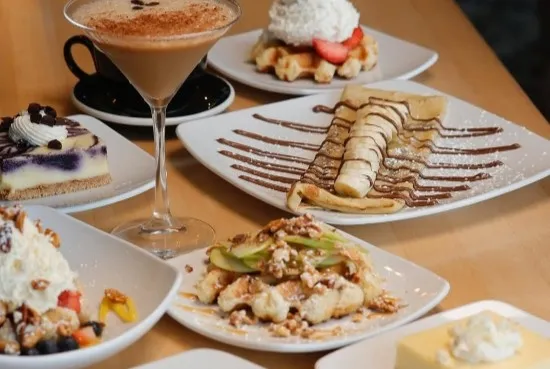 Spread of Taste of Belgium desserts including a Liège waffle with strawberries, a chocolate-drizzled crêpe, cheesecake and an espresso martini on a wood table