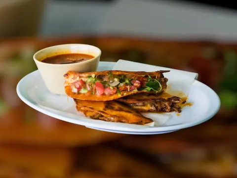 Quesabirria taco with melted cheese and pico de gallo served with a cup of consommé for dipping at Morelia's Taqueria