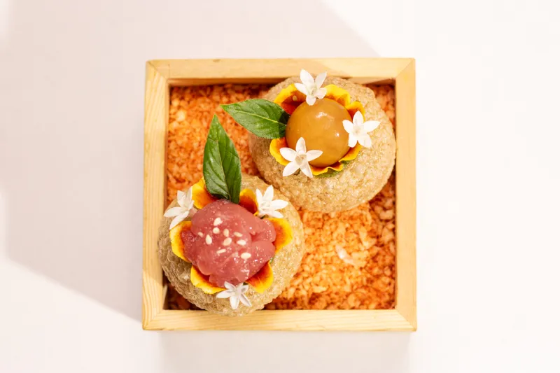 Two crisp pani puri-style bites topped with tuna and a curry pearl, garnished with basil and edible flowers, served in a wooden box at Amrina