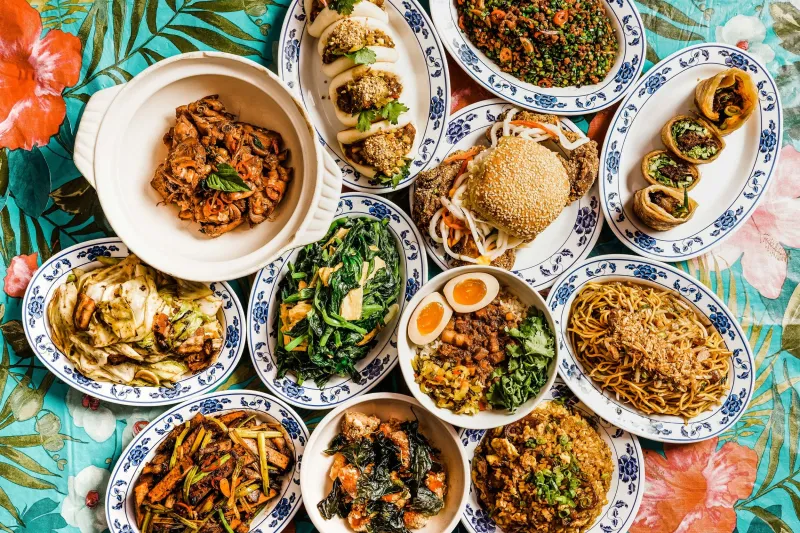 An overhead spread of 886's Taiwanese dishes in New York — gua bao, three-cup chicken, fly's head, lo ba beng, noodles and stir-fried greens on blue-and-white porcelain plates over a floral tablecloth