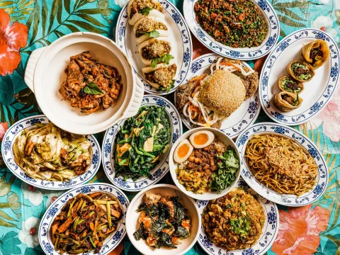 An overhead spread of 886's Taiwanese dishes in New York — gua bao, three-cup chicken, fly's head, lo ba beng, noodles and stir-fried greens on blue-and-white porcelain plates over a floral tablecloth