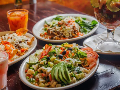 Spread of Polvos dishes on a dark wood table — a plate of ceviche and nopales salad with sliced avocado, tostadas, a shrimp cocktail in a glass, and aguas frescas
