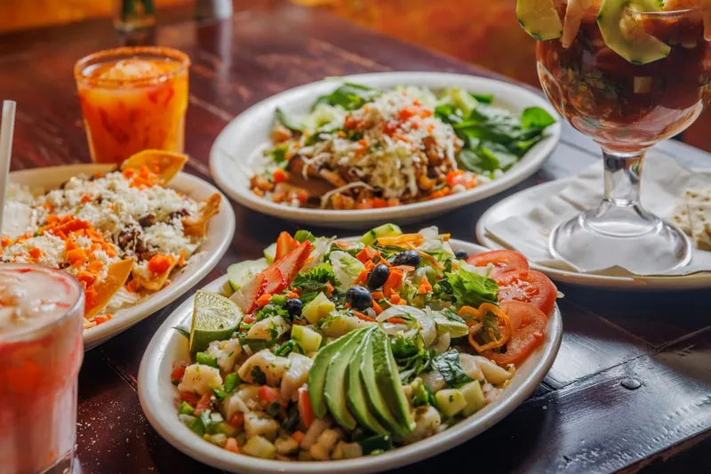 Spread of Polvos dishes on a dark wood table — a plate of ceviche and nopales salad with sliced avocado, tostadas, a shrimp cocktail in a glass, and aguas frescas