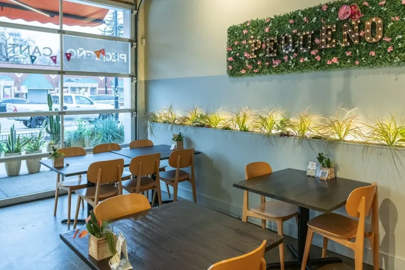 The dining room at Pequeño Cantina, with wooden tables and chairs beneath an illuminated "PEQUEÑO" marquee sign set into a greenery wall