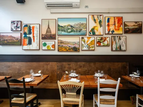 Dining room at Spinasse in Seattle, with set wooden tables and a bench banquette below a wall of framed Piedmont landscape and abstract art