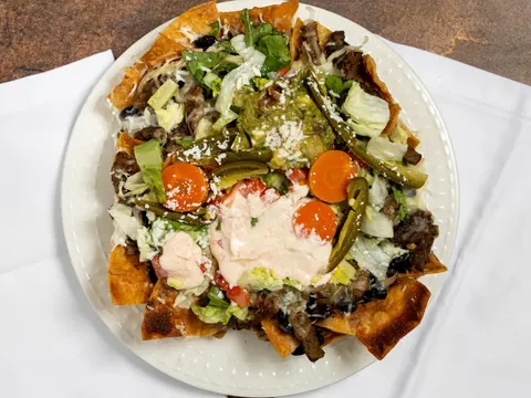 An overhead shot of loaded nachos at Real Taqueria topped with steak, guacamole, chipotle crema, cotija cheese, and pickled jalapeños and carrots on a white plate