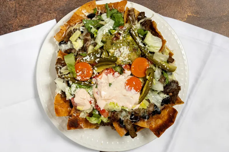 An overhead shot of loaded nachos at Real Taqueria topped with steak, guacamole, chipotle crema, cotija cheese, and pickled jalapeños and carrots on a white plate