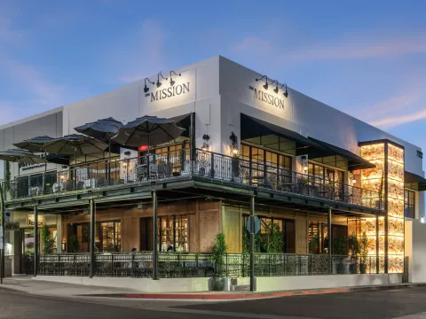 The Mission Old Town restaurant on a Scottsdale corner at dusk, with its illuminated onyx wall, wrought-iron balcony, patio umbrellas and lit 'The Mission' signage