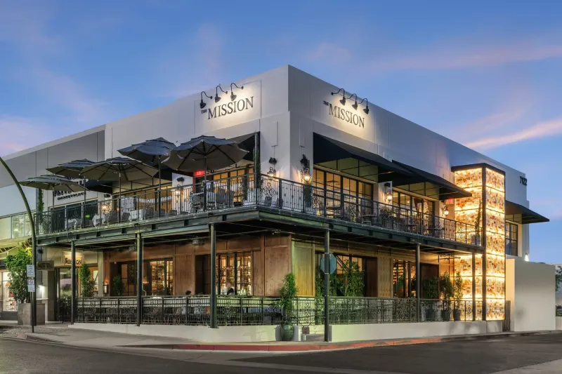 The Mission Old Town restaurant on a Scottsdale corner at dusk, with its illuminated onyx wall, wrought-iron balcony, patio umbrellas and lit 'The Mission' signage