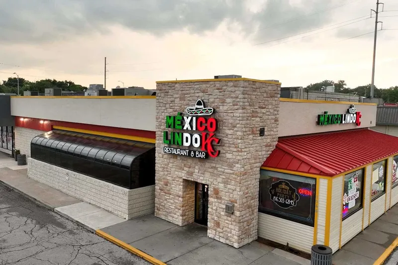 Exterior of Mexico Lindo KC Restaurant & Bar at 1046 W 103rd St, Kansas City, showing the stone facade, red roof and lit 'México Lindo KC Restaurant & Bar' sign with a sombrero logo