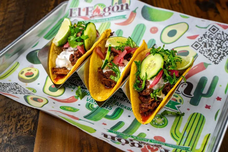 Three El Fogón Taquería street tacos on a branded paper-lined metal tray, topped with cilantro, cucumber and pickled onion, with lime wedges
