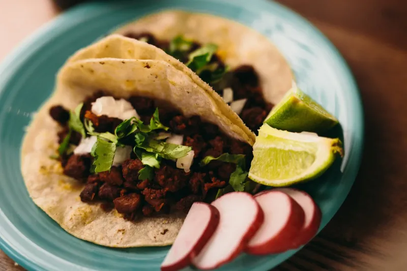 Two chorizo street tacos on soft corn tortillas topped with cilantro and onion, with lime wedges and sliced radish on a turquoise plate, at Boca Chica in Saint Paul