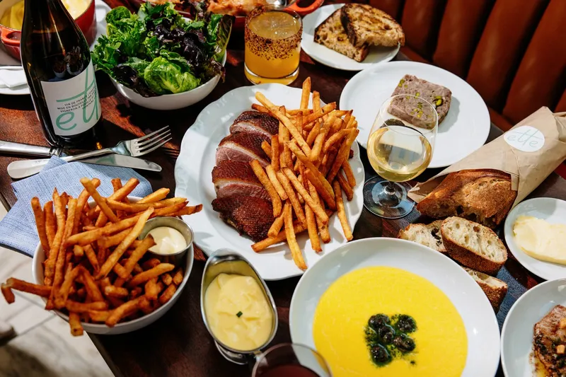 Table spread at Frenchette in Tribeca, New York with sliced duck and pommes frites, a green salad, country pâté, grilled bread and a glass of white wine