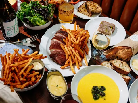 Table spread at Frenchette in Tribeca, New York with sliced duck and pommes frites, a green salad, country pâté, grilled bread and a glass of white wine