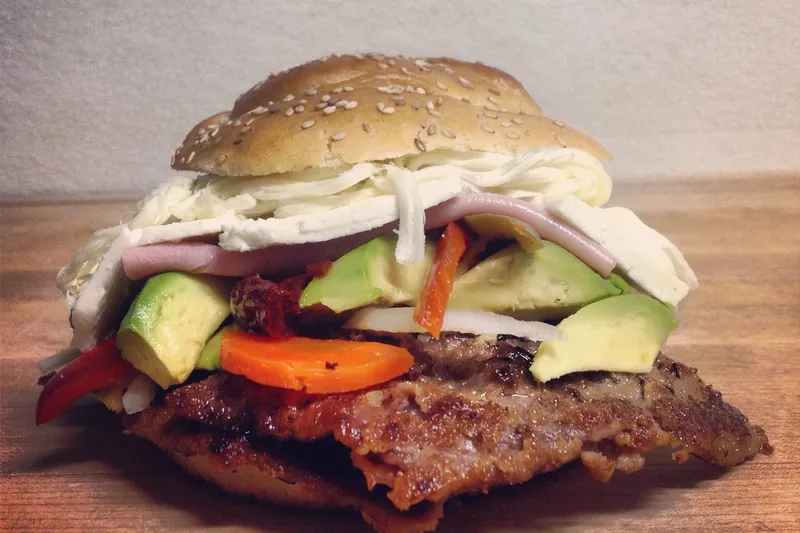 A cemita poblana from La China Poblana in East Los Angeles — a sesame-seed roll layered with a breaded beef milanesa, avocado, pickled carrots and jalapeños, onion, and shredded and sliced white cheese, on a wooden board