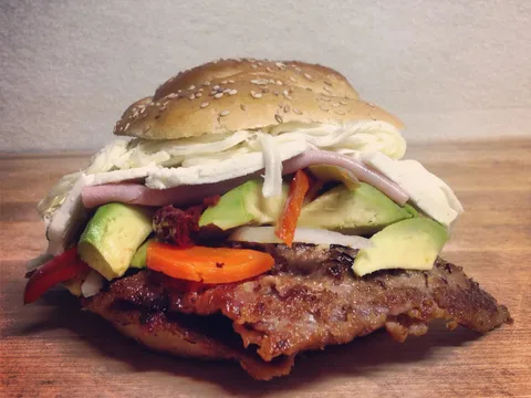 A cemita poblana from La China Poblana in East Los Angeles — a sesame-seed roll layered with a breaded beef milanesa, avocado, pickled carrots and jalapeños, onion, and shredded and sliced white cheese, on a wooden board