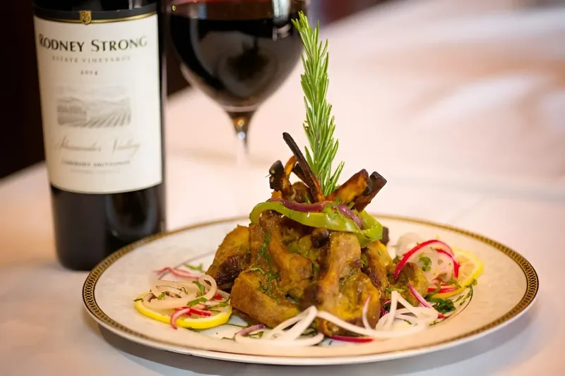 A plated dish of tandoori lamb chops garnished with rosemary, sliced onion, radish and lemon beside a glass of red wine at Narin's Bombay Brasserie