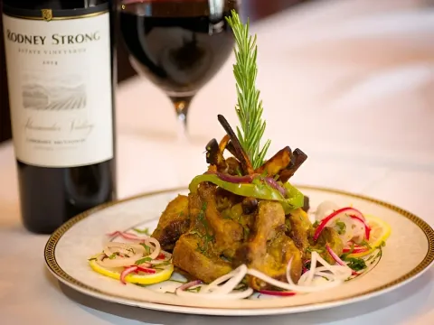 A plated dish of tandoori lamb chops garnished with rosemary, sliced onion, radish and lemon beside a glass of red wine at Narin's Bombay Brasserie