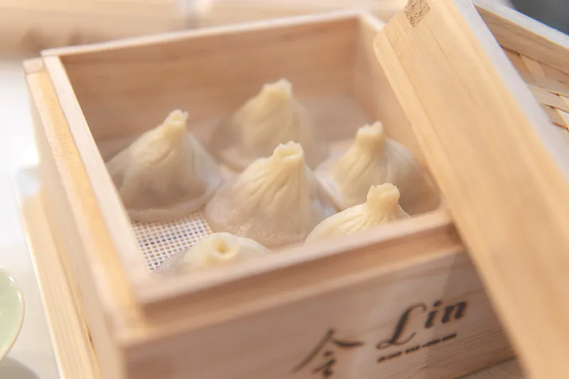 A bamboo steamer of xiao long bao soup dumplings branded with the restaurant's own '令 Lin' logo and 'Asian Bar + Dim Sum' mark, served at Lin Asian Bar + Dim Sum at 1203 W 6th Street in Austin