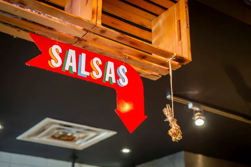 Interior detail at Resident Taqueria in Dallas: a red hand-painted 'Salsas' arrow sign hanging beneath a wooden crate shelf, with a bundle of dried chiles alongside