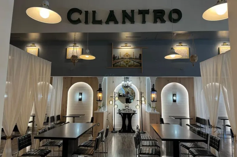 Interior dining room of Cilantro India in Cary, with large green CILANTRO lettering on the wall above a framed Taj Mahal photo, pendant lights, and black high-top tables with curtain dividers