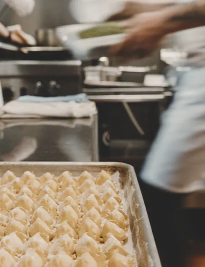 A sheet tray of freshly made, flour-dusted pasta in the kitchen at Dante in Omaha, with a cook working at the stove behind
