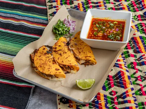 An order of Taqueria Number 1 quesabirria tacos — crispy cheese-filled tacos with a cup of red beef consomé, chopped onion and cilantro, on a paper-lined tray