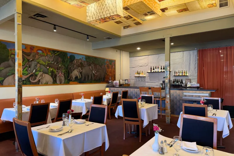 Ajanta's dining room in Berkeley with white-clothed tables, a long elephant mural, and a wine bar under a gilded panel ceiling