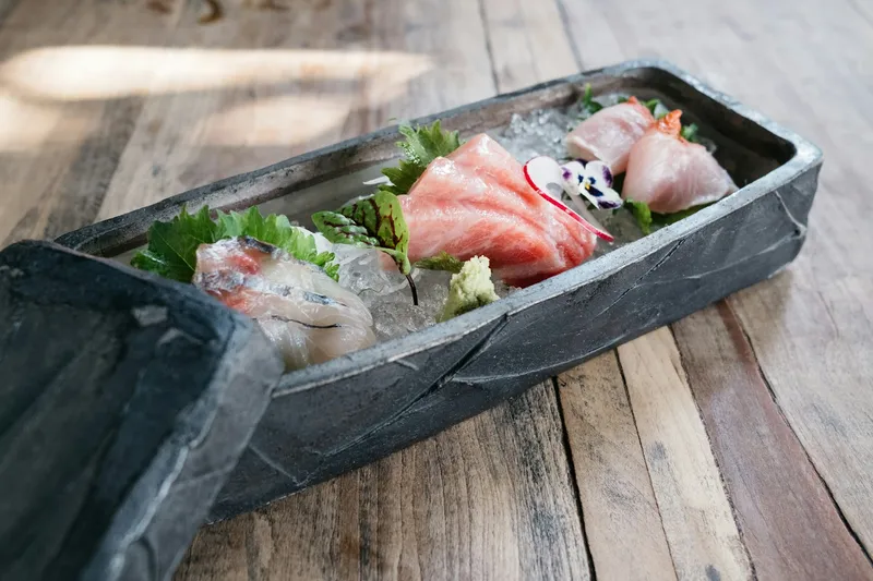Assorted sashimi over crushed ice, served in a dark stone boat-shaped dish on a wooden table at Kata Robata in Houston