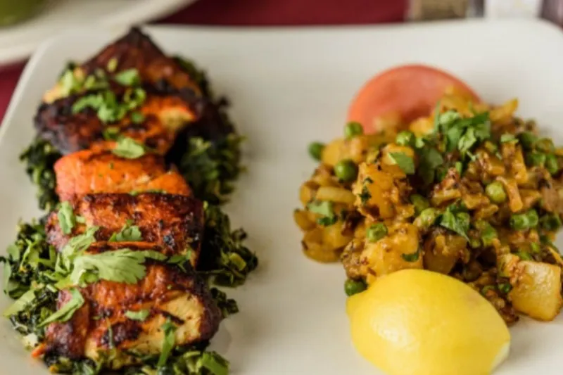 A white plate with charred tandoori chicken over sauteed greens beside a spiced potato-and-pea side and lemon at Silver Coin Indian Grill in Birmingham