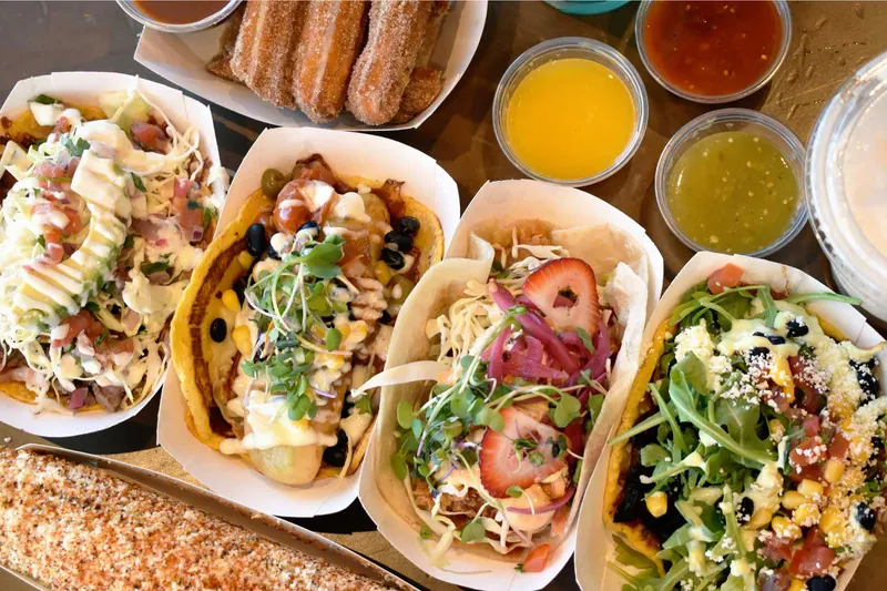 Four gourmet City Tacos in paper boats topped with microgreens, strawberry, arugula and cotija, alongside churros, salsas and elote