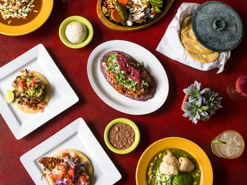 Overhead spread of tacos, a huarache, pozole verde, rice and beans on a red table at El Comal