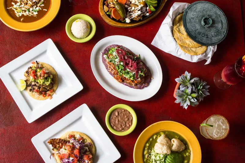 Overhead spread of tacos, a huarache, pozole verde, rice and beans on a red table at El Comal
