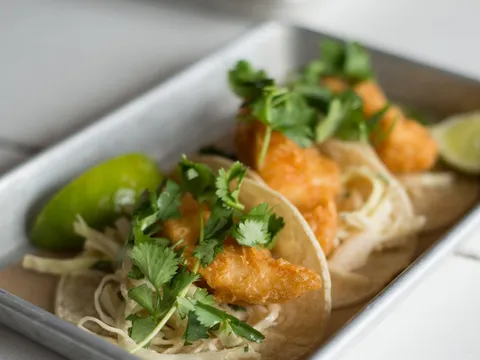 Crispy battered shrimp tacos topped with cilantro, cabbage slaw and lime wedges on a metal tray at Bartaco